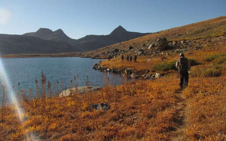 A group of people walk on a trail alongside a blue alpine lake. There are mountains in the background. 